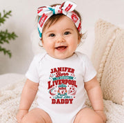 A smiling baby wearing a white short-sleeve bodysuit with a red and teal graphic that reads 'Janifer Born to Support Liverpool Just like Daddy' and a matching patterned bow headband, seated on a cream knit cushion.