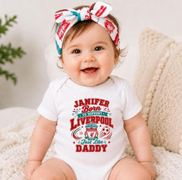 A smiling baby wearing a white short-sleeve bodysuit with a red and teal graphic that reads 'Janifer Born to Support Liverpool Just like Daddy' and a matching patterned bow headband, seated on a cream knit cushion.