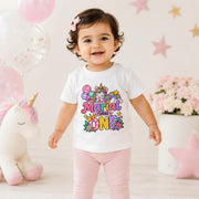 A smiling toddler stands in a pastel party setting wearing a white short-sleeve T-shirt with a colorful unicorn graphic and the words 'Marial is turning ONE' in pink, purple, yellow, and blue lettering. The child is also wearing light pink pants, with balloons, a plush unicorn, flowers, and star decorations in the background.