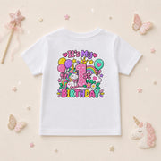 A white short-sleeve kids' t-shirt with a colorful 'It's My 1st Birthday' unicorn graphic, displayed on a light beige background with decorative pastel props including butterflies, a wand, and a unicorn plush.