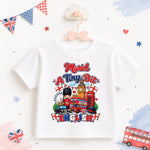 A white kids' short-sleeve T-shirt hanging on a pink bow hanger, featuring a colorful graphic print with London-themed elements and the text 'Murd A Tiny Bit ENGLISH'. The background is decorated with British-themed paper flags, stars, and a small red double-decker bus.