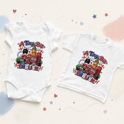 A white baby bodysuit and a white toddler T-shirt laid flat on a light cream background with pastel paint strokes and small star accents. Both garments feature a colorful graphic print reading “A Tiny Bit of ENGLAND” with a cartoon bear, red double-decker bus, London Eye, and London flags.