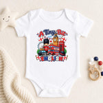 A white short-sleeve baby bodysuit laid flat on a cream fabric background, featuring a colorful graphic that reads 'A Tiny Bit ENGLISH' with London-themed illustrations including a red double-decker bus, red telephone booth, Big Ben, a bear, and English flags. A star toy, knit blanket, and wooden beads are placed around it as props.