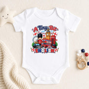 A white short-sleeve baby bodysuit laid flat on a cream fabric background, featuring a colorful graphic that reads 'A Tiny Bit ENGLISH' with London-themed illustrations including a red double-decker bus, red telephone booth, Big Ben, a bear, and English flags. A star toy, knit blanket, and wooden beads are placed around it as props.