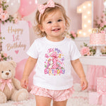 A toddler girl wearing a white short-sleeve t-shirt with a colorful 'It's My 1st Birthday' unicorn graphic and pink ruffled shorts, standing in a pink first birthday party setting.