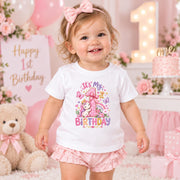 A toddler girl wearing a white short-sleeve t-shirt with a colorful 'It's My 1st Birthday' unicorn graphic and pink ruffled shorts, standing in a pink first birthday party setting.