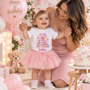 A toddler wearing a white graphic t-shirt and pink tutu skirt stands next to a woman in a pink dress at a baby shower or first birthday celebration. The scene is decorated with pink balloons, a teddy bear, and a cake.