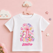 A white short-sleeve baby t-shirt with a colorful first birthday unicorn graphic, hanging on a pink hanger against a soft pink background with decorative cloud and star props.