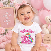 Smiling baby wearing a white personalised 10 months old unicorn bodysuit in a pink milestone celebration setting.