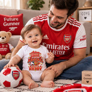 A man and a baby sit together in a living room, both wearing Arsenal-themed apparel. The man wears a red Arsenal home jersey, and the baby wears a white bodysuit with a red and gold Arsenal graphic that reads 'Adam Bean TO SUPPORT ARSENAL AND BE DADDY'.