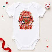 A white short-sleeve baby bodysuit laid flat on a light beige background, featuring a colorful graphic that reads 'Adam Born to Support Arsenal Just like Daddy' with the Arsenal FC crest and soccer ball motif. Decorative red ribbon, star confetti, and a small soccer ball are placed around the product.