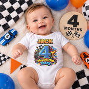 Smiling baby wearing a personalised 4 months old racing bodysuit beside toy cars and a wooden month marker.