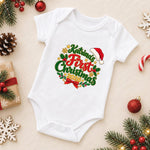 A white short-sleeve baby bodysuit laid flat on a light beige background, decorated with a colorful 'Katrina's First Christmas 2020' graphic. The bodysuit is surrounded by festive props including pine branches, red ornaments, a candy cane, a wrapped gift, and wooden snowflakes.