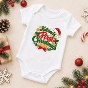 A white short-sleeve baby bodysuit laid flat on a light beige background, decorated with a colorful 'Katrina's First Christmas 2020' graphic. The bodysuit is surrounded by festive props including pine branches, red ornaments, a candy cane, a wrapped gift, and wooden snowflakes.