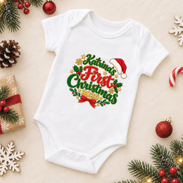 A white short-sleeve baby bodysuit laid flat on a light beige background, decorated with a colorful 'Katrina's First Christmas 2020' graphic. The bodysuit is surrounded by festive props including pine branches, red ornaments, a candy cane, a wrapped gift, and wooden snowflakes.