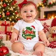 A baby sitting on a white shag rug in a cozy Christmas setting, wearing a white short-sleeve bodysuit with a festive graphic that reads 'My First Christmas 2024' in green, red, and gold. The baby is also wearing a red bow headband.