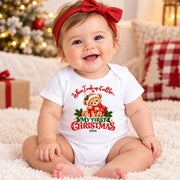 Smiling baby wearing a white personalised first Christmas teddy bear bodysuit and red bow beside a Christmas tree.