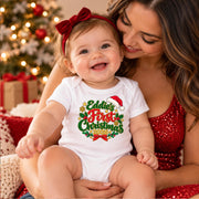 A baby wearing a white short-sleeve bodysuit with a colorful 'Eddie's First Christmas 2024' graphic sits on a woman's lap. The baby also wears a red bow headband.