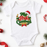 A white short-sleeve baby bodysuit laid flat on a white background, decorated with a colorful 'My First Christmas 2025' graphic featuring green and red lettering, a Santa hat, holly leaves, and gold stars. The bodysuit is styled with holiday props like candy canes, pine branches, ornaments, and a red ribbon.